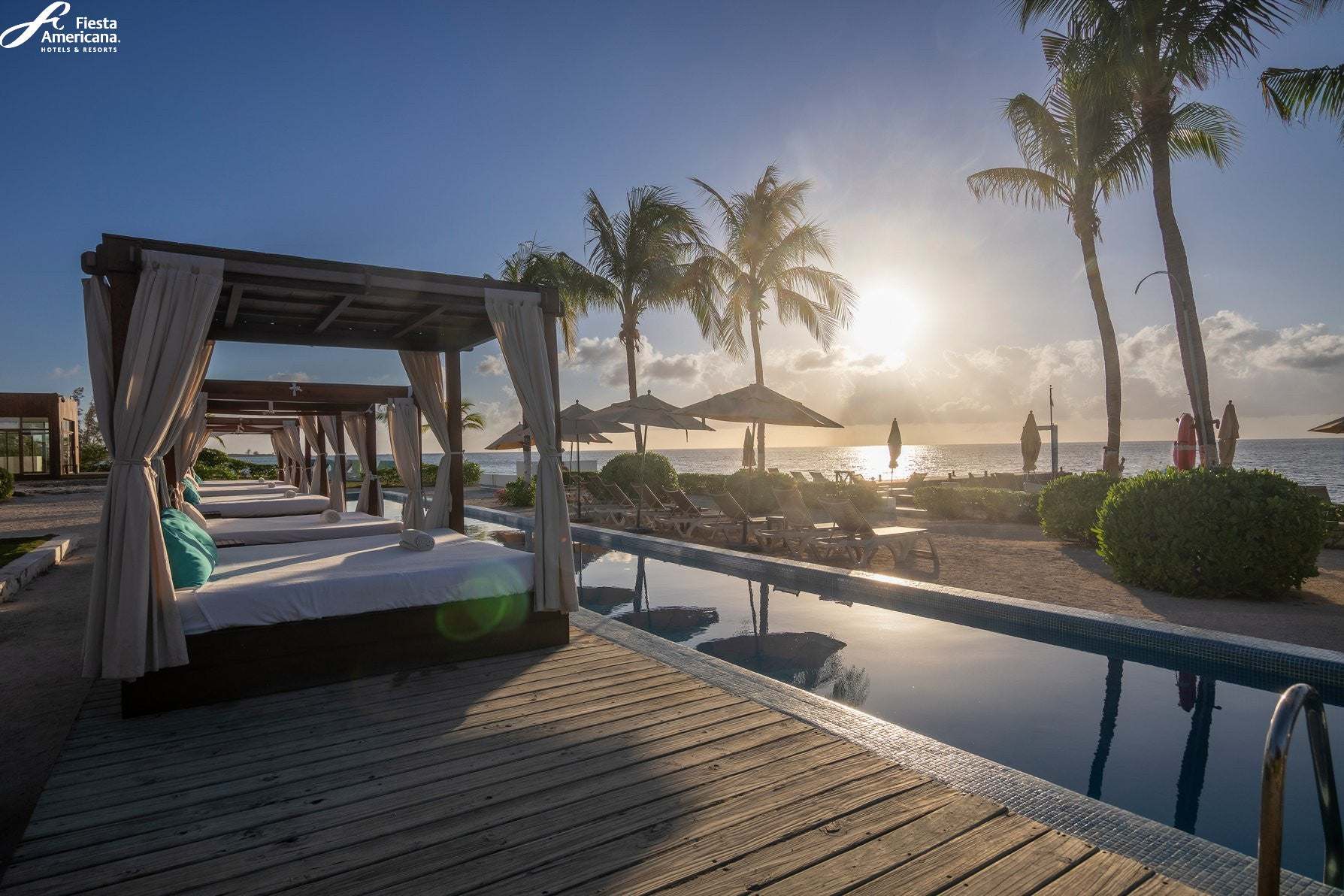Pool area with cabanas and palm trees at sunset, featuring the Fiesta Americana brand.